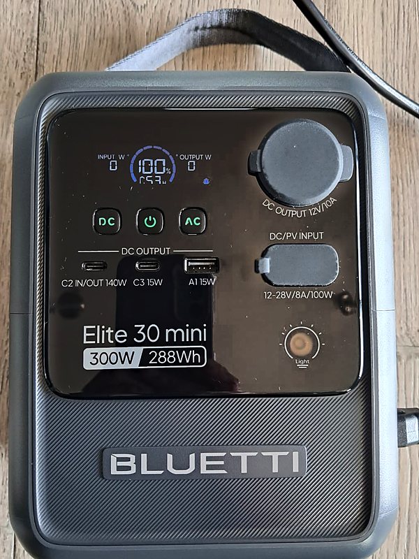 Our Bluetti Elite 30 Mini review unit on a wood floor, screen showing 100% charge, with its AC charging cable plugged in